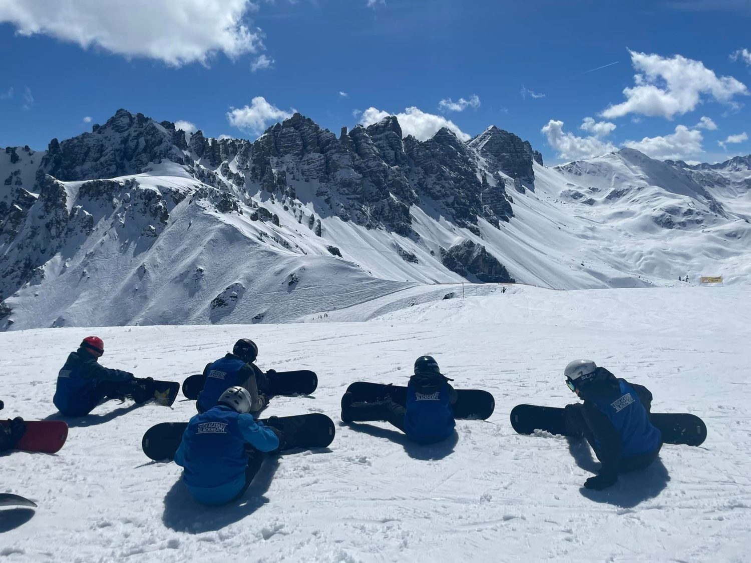 A group of LAB students are seen sitting on the snowy ground and looking at the mountains whilst on a Ski Trip to Austria.