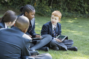 A group of four Year 7 students can be seen sitting on a grass area on the school grounds, using iPads.