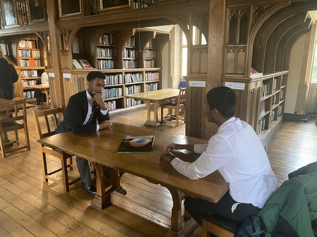 Two male students are seen sitting across from each other at a wooden table in the Library at Oxford University.