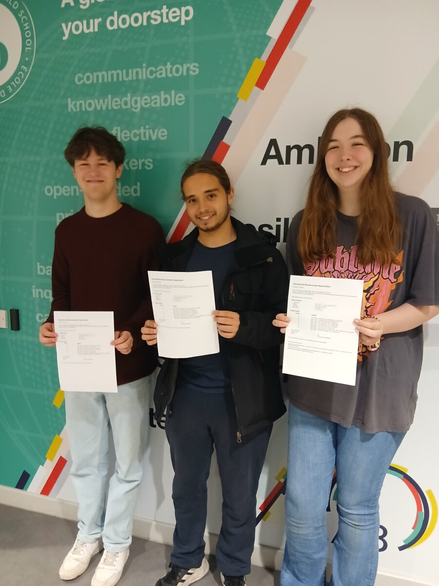 Three students are standing indoors, each holding papers. The background features text on a wall, with words like "communicators," "knowledgeable," and "reflective," which are often part of educational institution's values or mission statements. The papers they are holding appear to be official documents, consistent with receiving exam results.