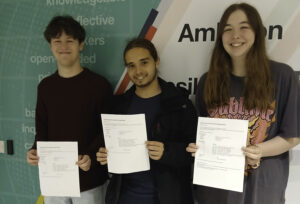 Three students are standing indoors, each holding papers. The background features text on a wall, with words like "communicators," "knowledgeable," and "reflective," which are often part of educational institution's values or mission statements. The papers they are holding appear to be official documents, consistent with receiving exam results.