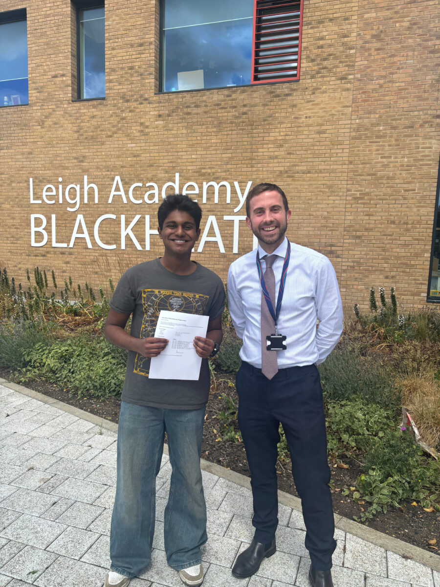 This photo shows a student with an adult male (likely a teacher or staff member) in front of a building with "Leigh Academy BLACKHEATH" visible on its facade. The students are holding papers, suggesting they are receiving their results or official documents. The bright outdoor setting and the papers held by the students indicate an important day, often associated with exam results.