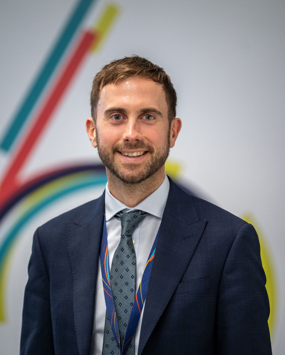 head-and-shoulders portrait of a smiling man with short brown hair and a beard. He is wearing a dark blue suit, a light blue shirt, a patterned tie, and a lanyard, standing in front of a white wall with a colorful, out-of-focus graphic.