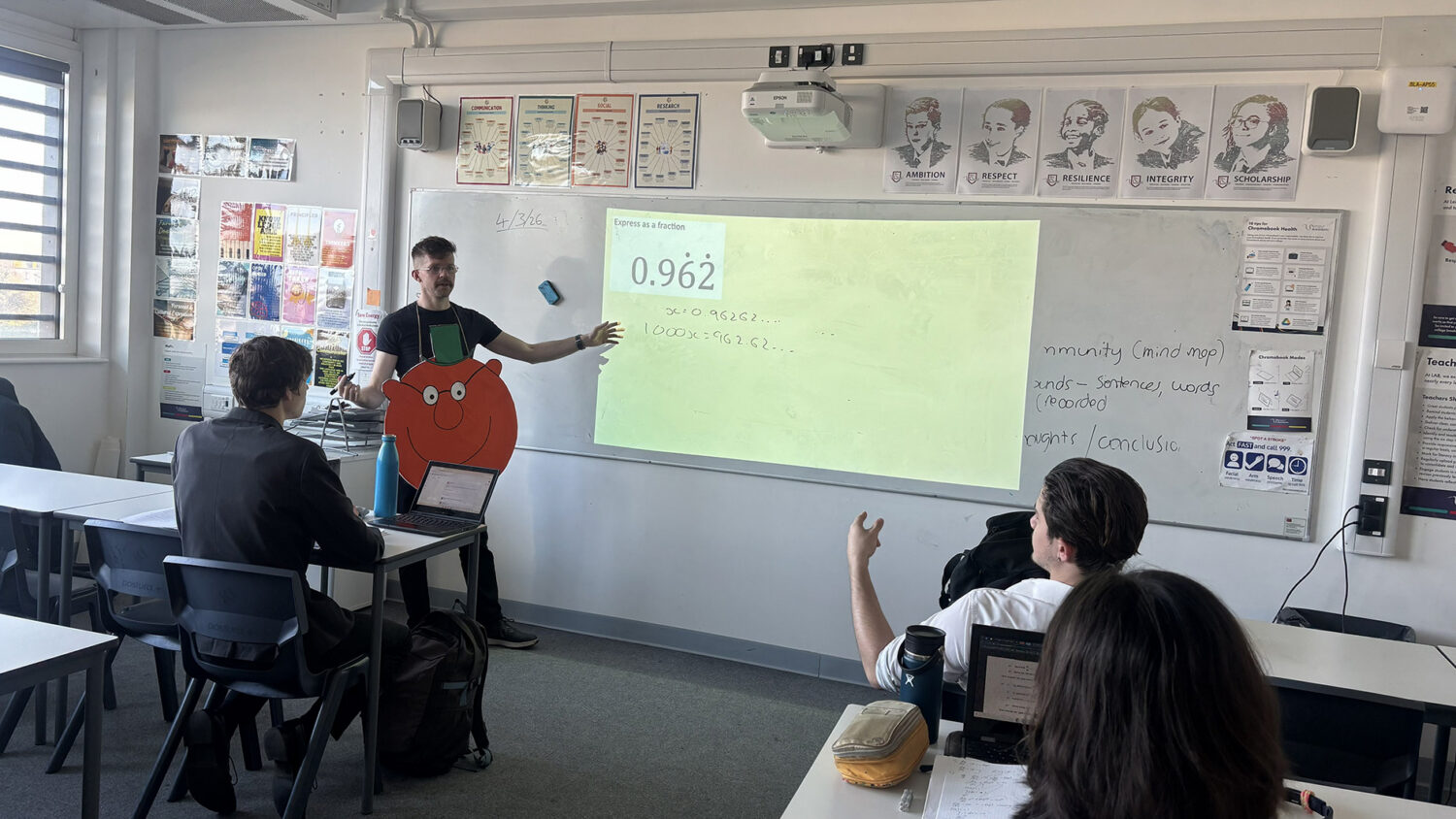 A teacher wearing a Mr. Men cardboard cutout around his neck teaches a math lesson using a projector in a classroom while students watch from their desks.