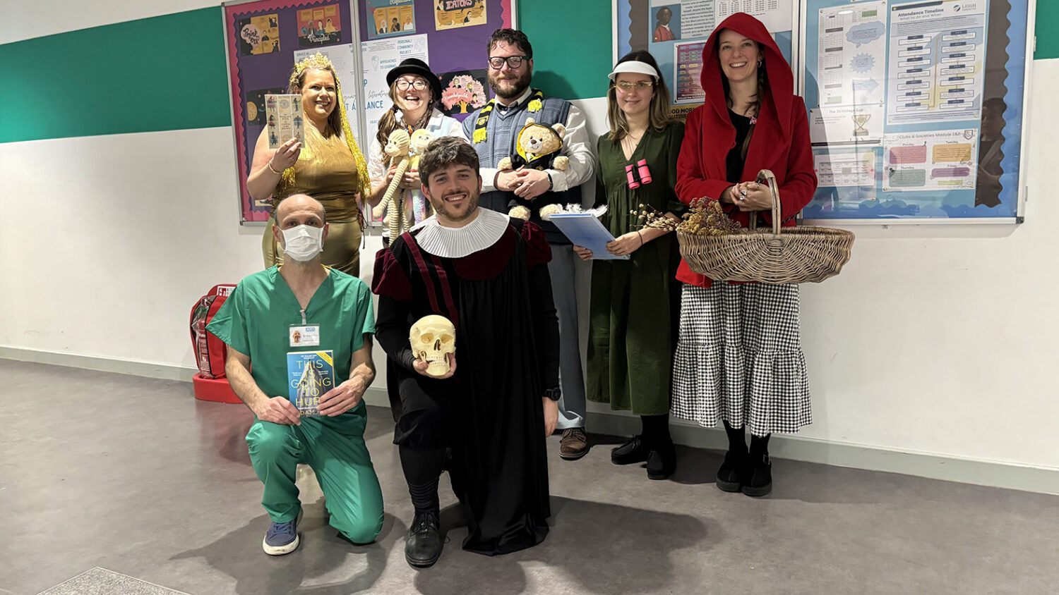 A group of six adults posing in a hallway, dressed in varied book character costumes, including a doctor holding a book, a person in a gold dress and crown, and someone in a Renaissance outfit holding a skull.