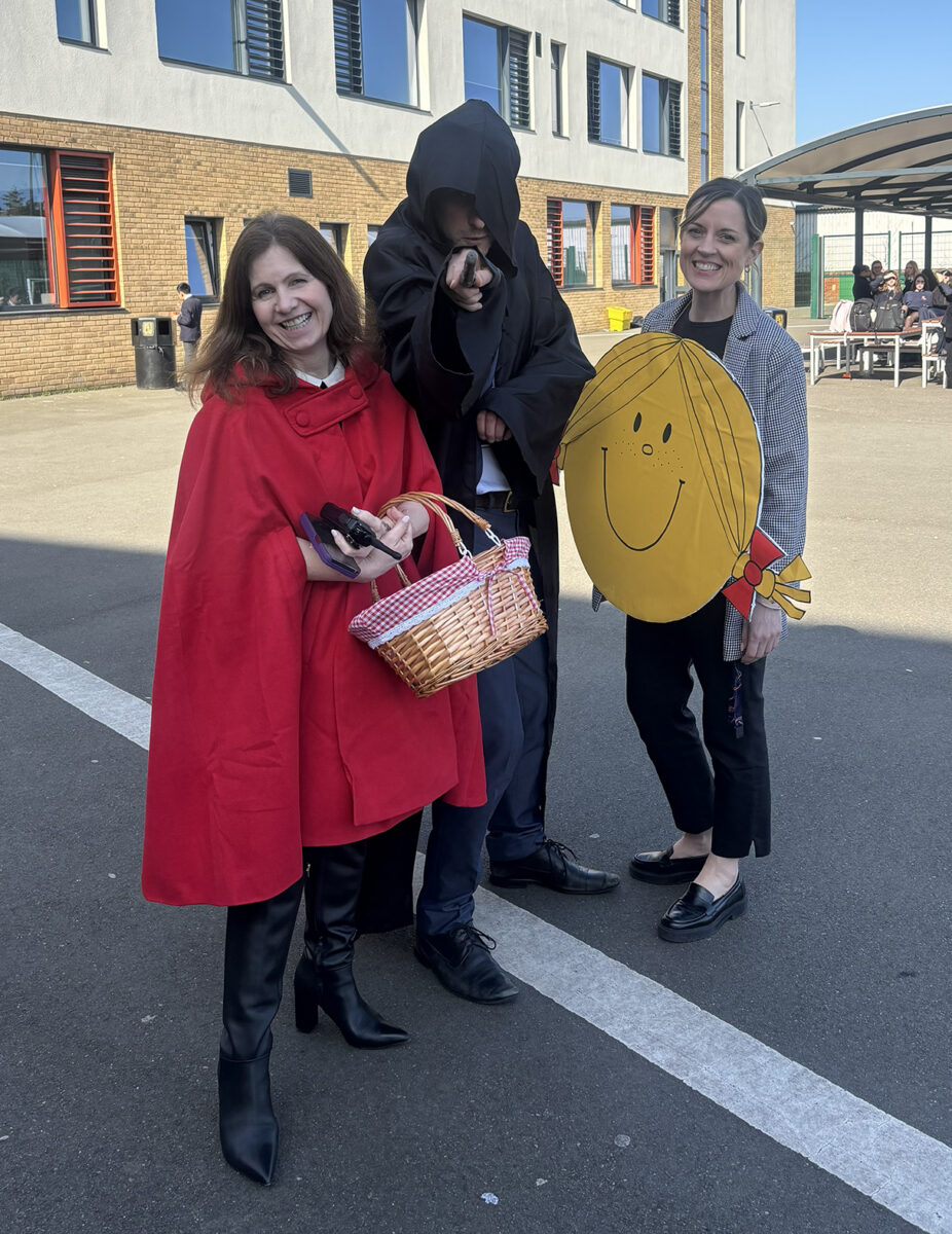 Three adults outdoors on a playground wearing costumes, featuring Little Red Riding Hood, a dark cloaked figure with a wand, and a Little Miss Sunshine cutout.