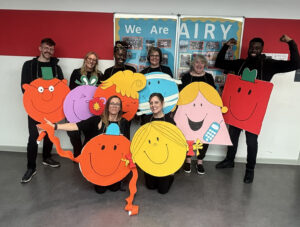 Seven adults smiling together indoors, all wearing large, colorful cardboard cutouts of Mr. Men and Little Miss characters over black clothing.