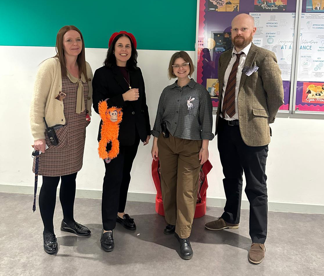 Four adults indoors dressed in character costumes, holding or wearing props like a stuffed orangutan and small paper animals pinned to their shirts.