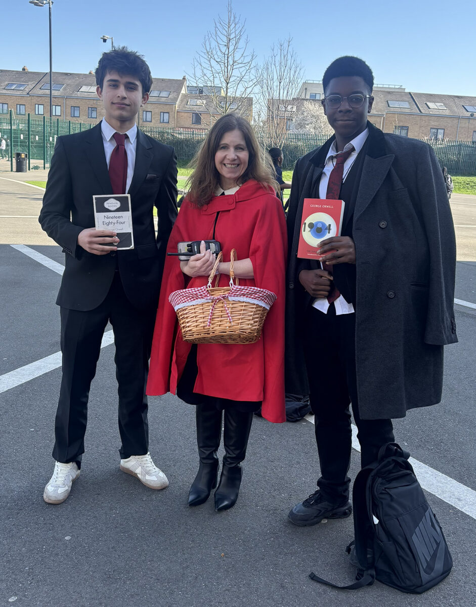 Two students in school uniforms holding copies of the book "1984," standing on either side of an adult dressed as Little Red Riding Hood outdoors.